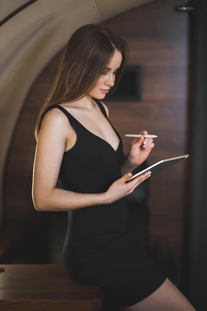 Lovely slender young woman wearing sexy black dress in private jet. Successful young woman entrepreneur holding digital tablet, working during the flight. Technology concept.