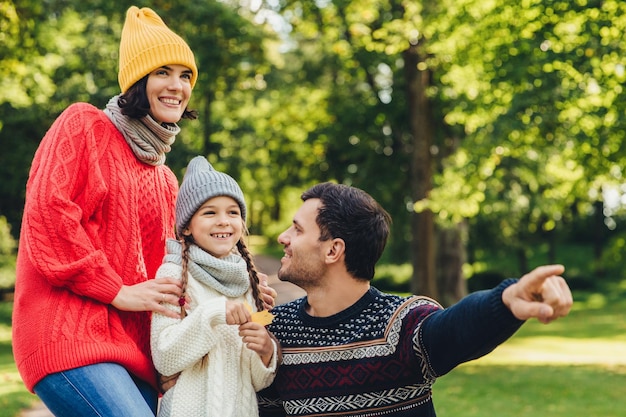 Lovely small girl with two pigtails wears knitted hat scarf and sweater holds leaf in hand looks joyfully into distance as her father shows her beautiful flower Spending autumn days together