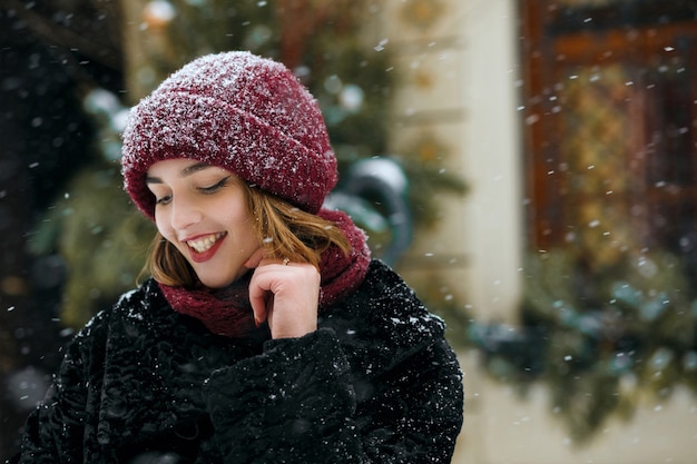 Lovely smiling brunette woman wearing red cap posing at the street during snowfall. Empty space