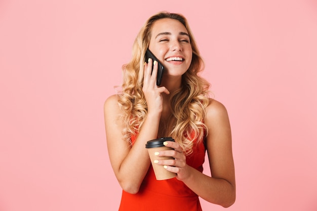 Foto bella sorridente giovane donna bionda che indossa abiti estivi in piedi isolato su un muro rosa, parlando al telefono cellulare mentre si tiene tazza da asporto