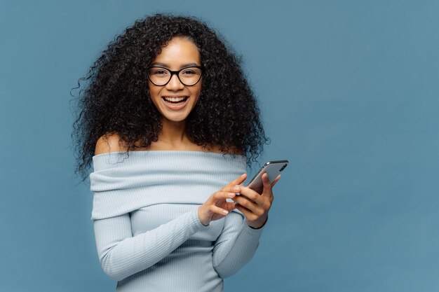 Lovely teenage girl enjoys online communication holds modern cell phone checks email box connected to wireless internet cannot imagine life without technologies isolated on blue studio wall