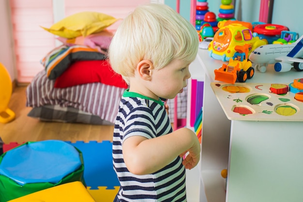 Lovely toddler boy in stripped tshirt standing near shelf with toys at the kindergarten looking away Little boy feeling insecure while his first day at kindergarten