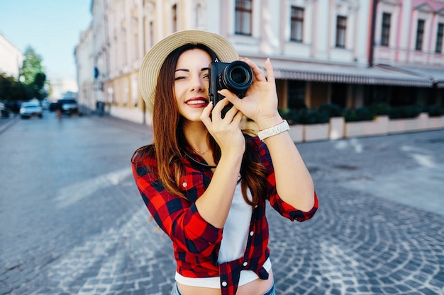 Bella ragazza turistica con capelli castani che indossa cappello e camicia rossa, facendo foto alla vecchia città europea e sorridente, viaggiando.