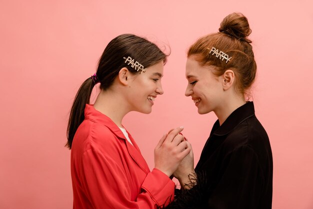 Lovely two caucasian young women brunette and redhead standing facing each other and holding hands on pink background Femininity and beauty sensuality concept