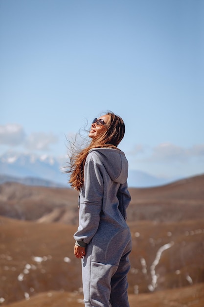 A lovely woman in a blue overalls and sunglasses is smiling happily and enjoying the mountain nature