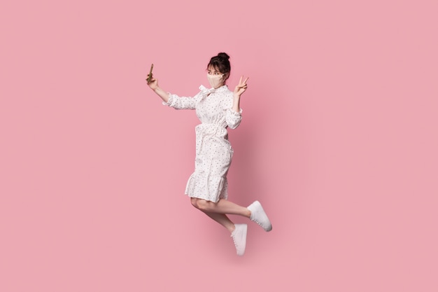 Lovely woman in a dress jumping and making a selfie on a pink studio wall while wearing a medical mask on face