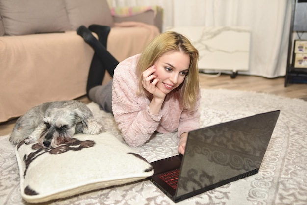 Прекрасная женщина в розовом свитере лежит на ковре с ноутбуком дома, серая собака спит на подушке рядом с ней