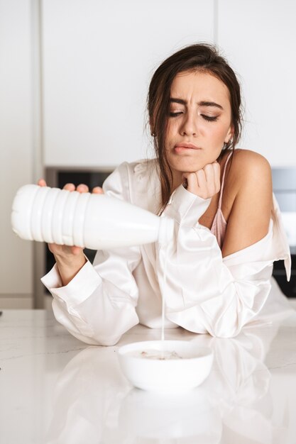 lovely woman wearing silk clothing eating granola with milk, while having breakfast in apartment