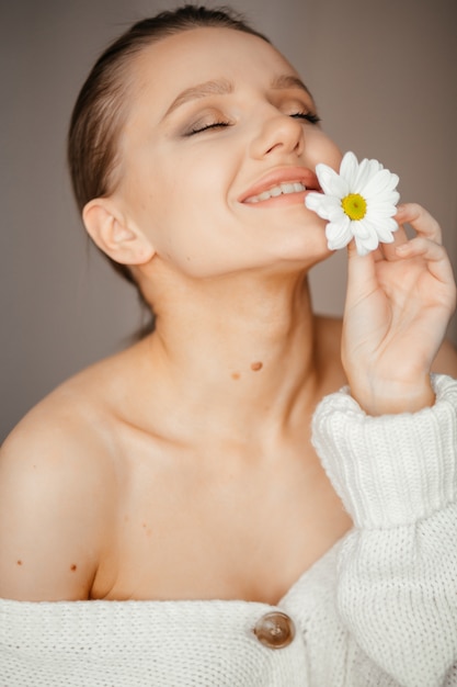 Lovely woman with closed eyes in a white sweater brings a white flower to her lips