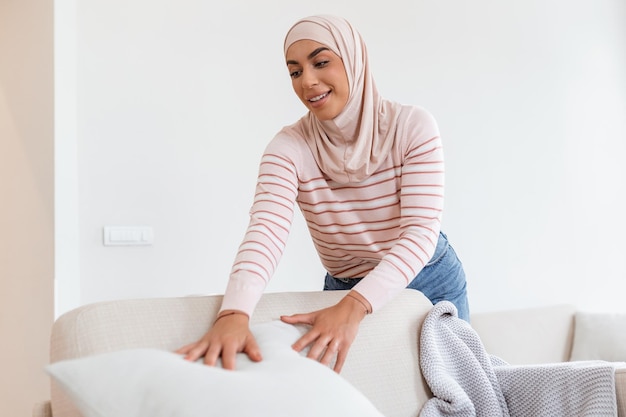 Lovely young arabic muslim woman putting soft pillows and plaid\
on comfy sofa making her home cozy and warm