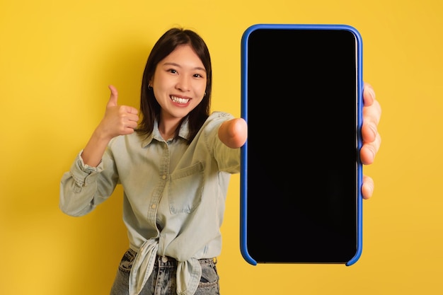 Lovely young asian woman demonstrating smartphone with mockup gesturing thumb up promoting