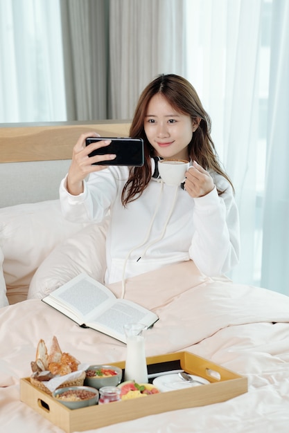 Lovely young Asian woman having breakfast in bed, she is taking selfie or filming video with cup of coffee in hand