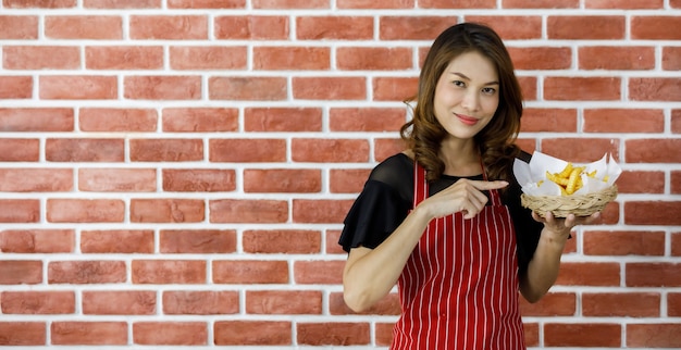 Lovely young Asian woman in red apron smilingly showing wicker basket of tasty fried potatoes between brick wall and counter of kitchenwares in home as proud and enjoy self cooking delicious food menu