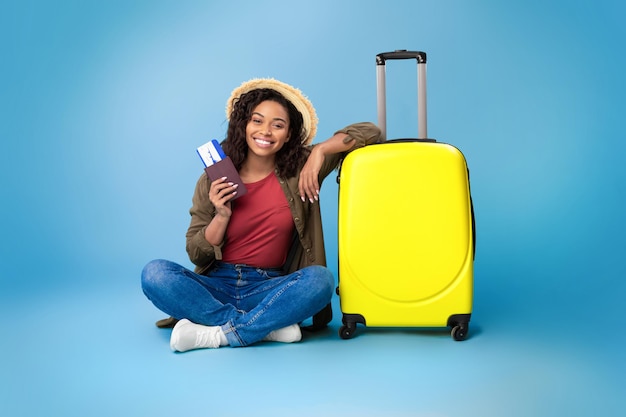 Lovely young black woman holding passport and plane tickets sitting near bright yellow suitcase on