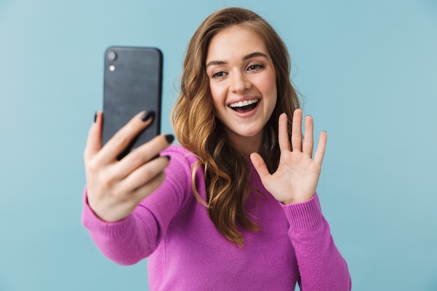 Bella giovane donna bionda in piedi isolata sul muro blu, facendo un selfie, agitando la mano