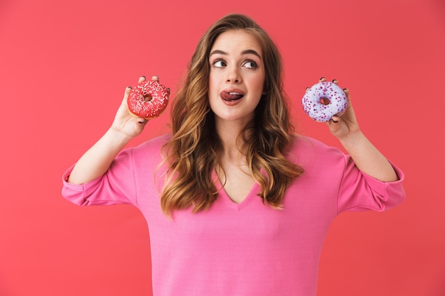 Lovely young blonde woman standing isolated over pink wall, holding donuts