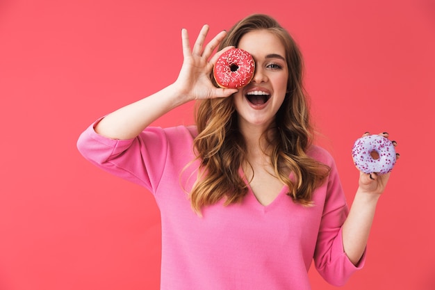 Lovely young blonde woman standing isolated over pink wall, holding donuts