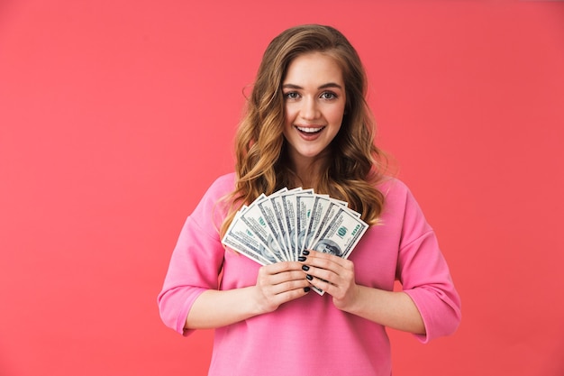 Lovely young blonde woman standing isolated over pink wall, showing money banknotes