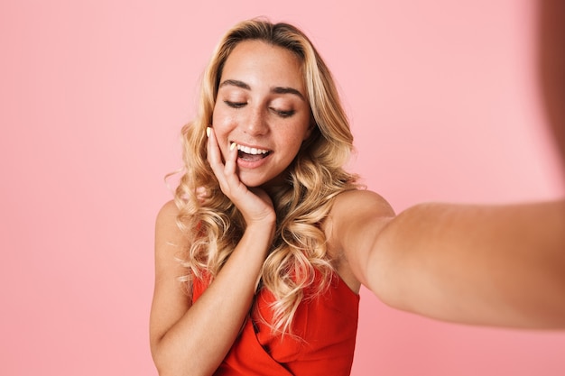 Lovely young blonde woman wearing summer dress standing isolated over pink wall, taking a selfie