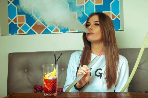 Lovely young brunette woman with green eyes smokes shisha and drinks cocktail