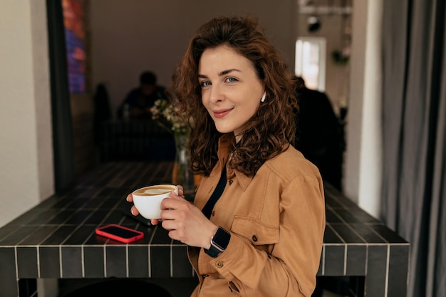 Lovely young caucasian girl looks into camera sitting at table and holding cup of coffee in cafe Brunette wearing brown shirt is working on project Programmer concepts