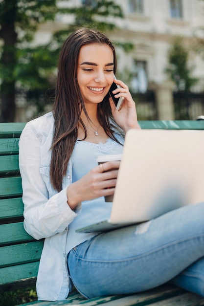 Lovely young female talking at her smartphone while looking at screen of a laptop while sitting on bench and drinking coffee outside.