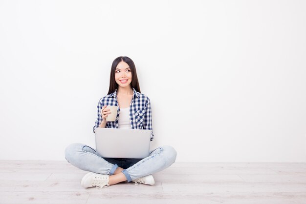 L'usura adorabile della ragazza in abiti casual è seduta sul pavimento che beve il pc della stretta del caffè