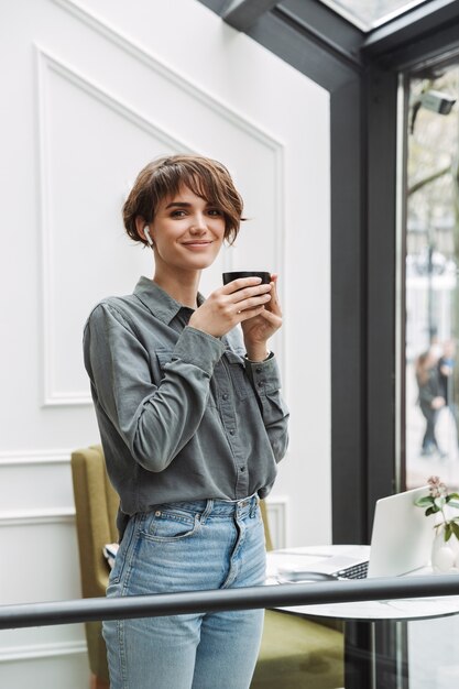 Lovely young girl wearing wireless earphones drinking coffee while standing at the cafe indoors