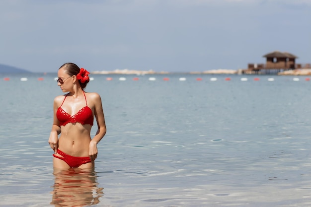 빨간 니트 수영복을 입은 사랑스러운 젊은 여성이 멀리 바라보고 바닷물 속을 걷고 있습니다.