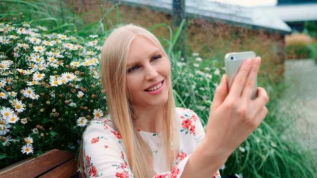 Lovely young lady using smartphone to make video call while sitting on bench on living roof of huge mall in usa