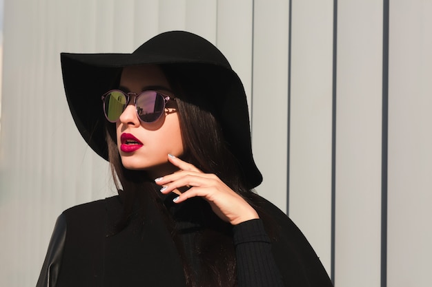 Lovely young model in mirror glasses and broad brimmed hat posing near the shutters