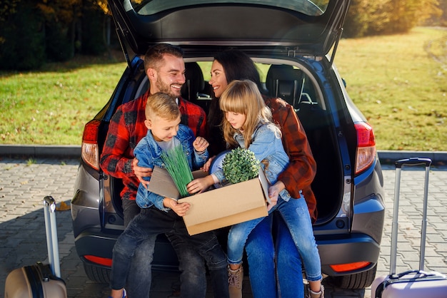 Lovely young parents with their cute kids sitting in the trunk and holding carton box with plants