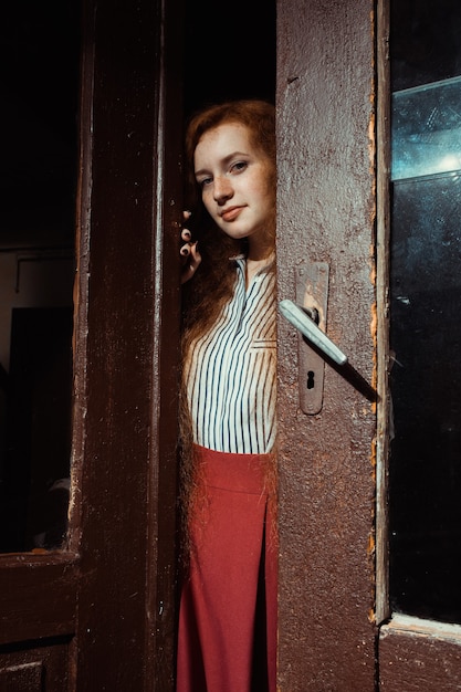 Милая молодая рыжеволосая модель с длинными волнистыми волосами открывает старую деревянную дверь