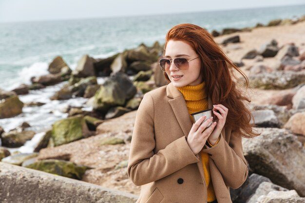 Lovely young redhead woman wearing autumn coat walking at the beach, drinking hot coffee from thermos