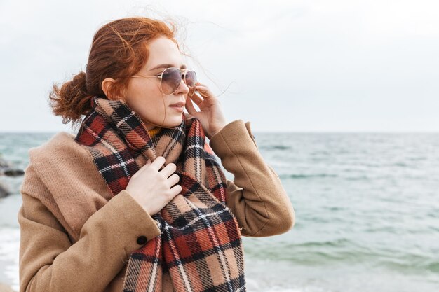 Lovely young redhead woman wearing autumn coat walking at the beach