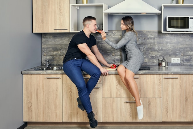 Lovely young white woman sits on an induction stove built into kitchen table and feeds an attractive  man with sweet pepper.