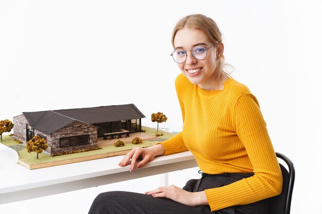 Photo lovely young woman architect sitting at the desk with house model isolated over white wall