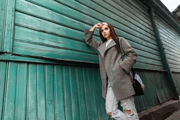 Bella giovane donna in bandana alla moda in cappotto vintage in jeans strappati con borsa in pelle posa vicino a edificio verde da assi. attraente ragazza graziosa affascinante in abiti alla moda all'aperto.