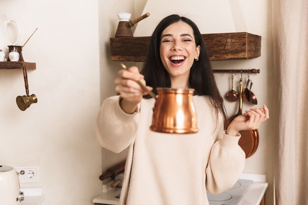 Lovely young woman having cup of coffee at the kitchen, showing coffee maker