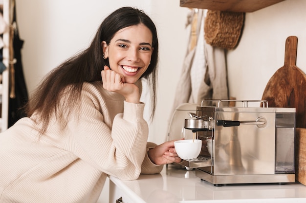 Прекрасная молодая женщина с чашкой кофе на кухне, используя кофеварку