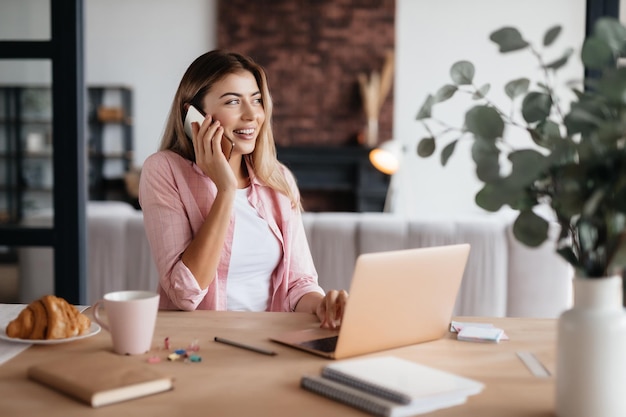 Lovely young woman looking pleased while talking on the phone and sitting at the table with laptop