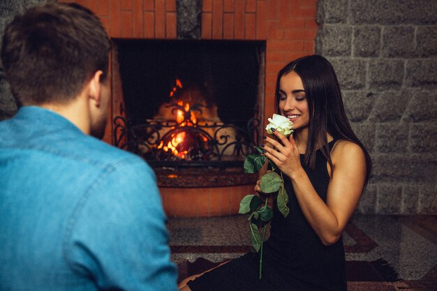 Lovely young woman smell white rose. She hold it close to face and look at it Guy sit with back to camera. They are near fireplace.