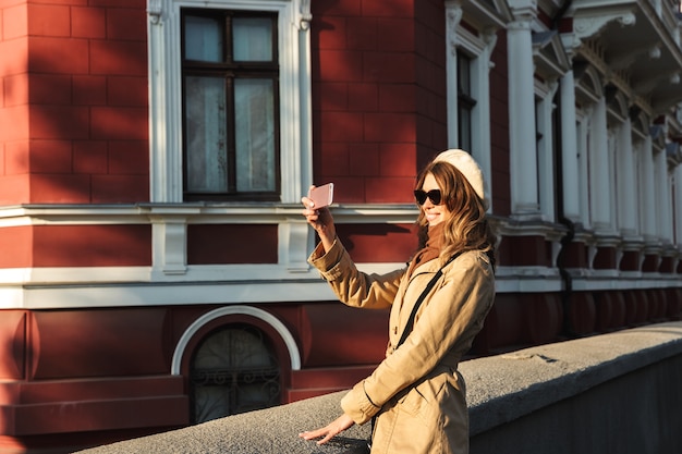 Foto bella giovane donna che indossa un cappotto che cammina all'aperto, prendendo un selfie