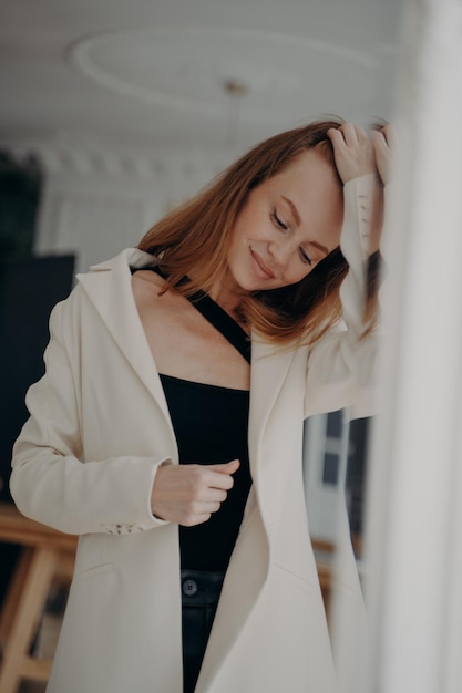 Lovely young woman wearing white suit jacket fixes hair posing near mirror Female fashion beauty