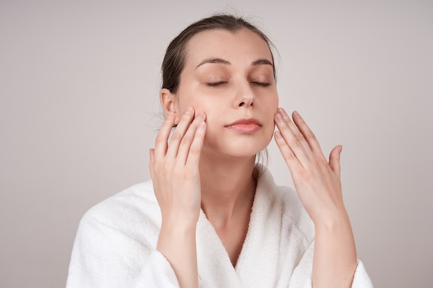 A lovely young woman in a white robe, touching her cheeks, enjoying the softness of her skin. Facial care concept.