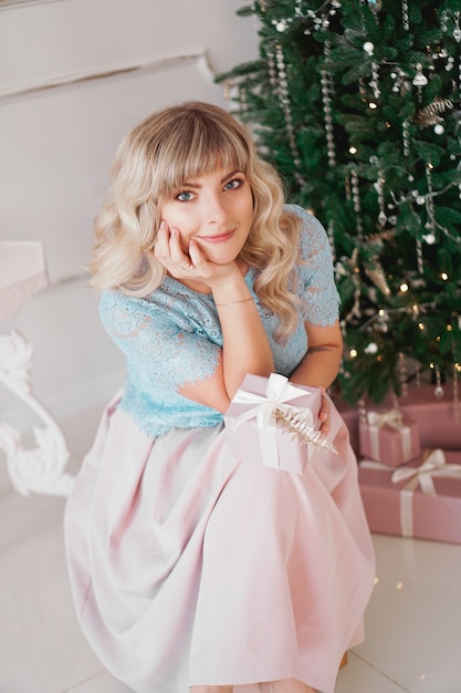 ピンクのクリスマスプレゼントで飾られた木の近くに屋内に座っているエレガントなスタイルの素敵な若い女性