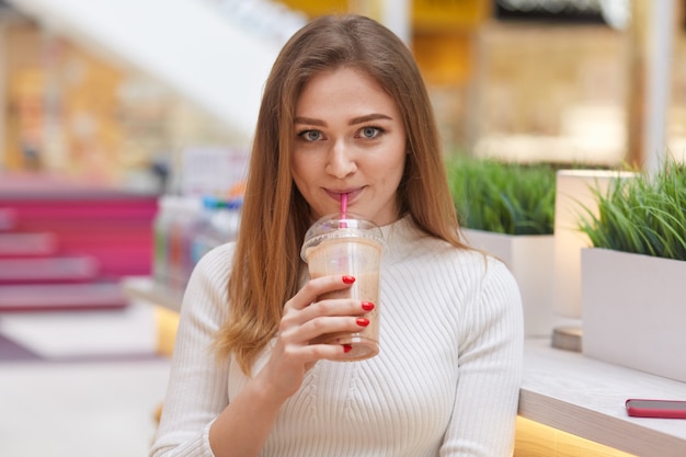 Lovely young woman with satisfied expression, drinks milk cocktail, wears white jumper, has make up