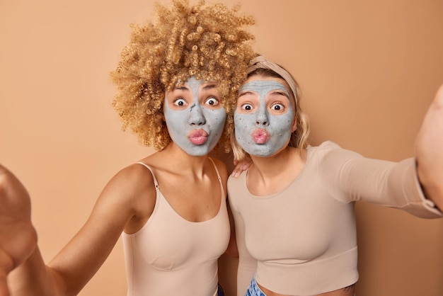 Lovely young women make grimace at camera keep lips folded apply beauty clay mask to reduce fine lines dressed casually isolated over brown background Facial treatments and skin care concept