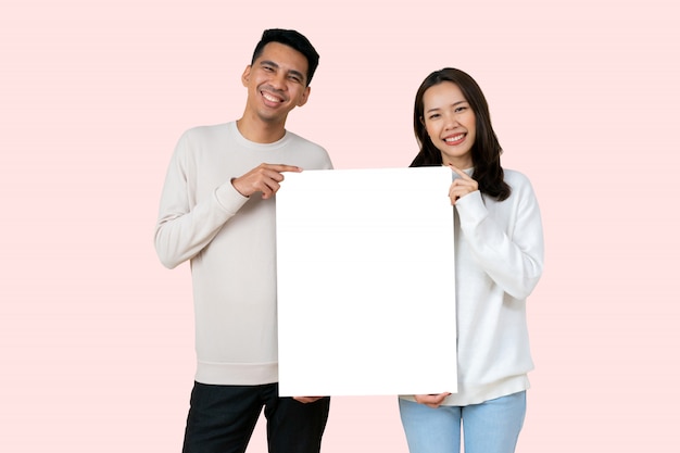 恋人のアジア人はバレンタインの日にピンク色の背景に分離された白いモックアップボードを一緒に保持します。