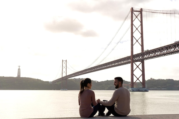 リスボンの4月25日橋を背景にテージョ川のほとりでお互いを見つめる恋人たち